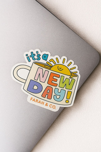 Colorful “It’s a New Day” sticker featuring a smiling sun inside a mug with playful pastel text, placed on a silver laptop – by Farah & Co.